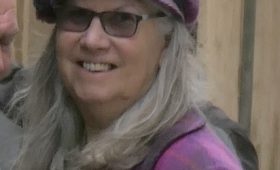 women with mid long grey hair wearing a purple cap and coat, smiling. Wearing sunglasses