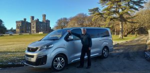 man standing in front of a grey van. less castle is visible in the background